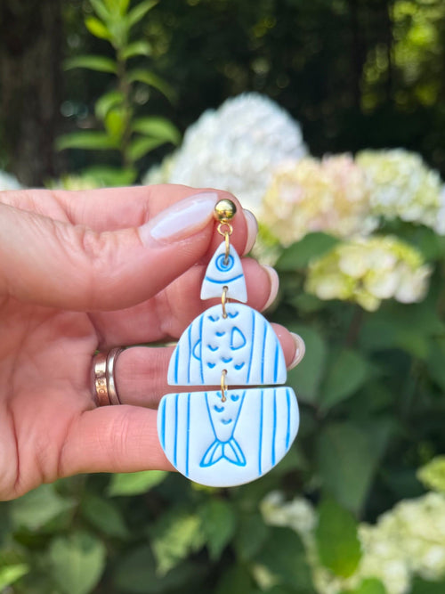 Handmade blue fish dangle earrings held up against a garden background